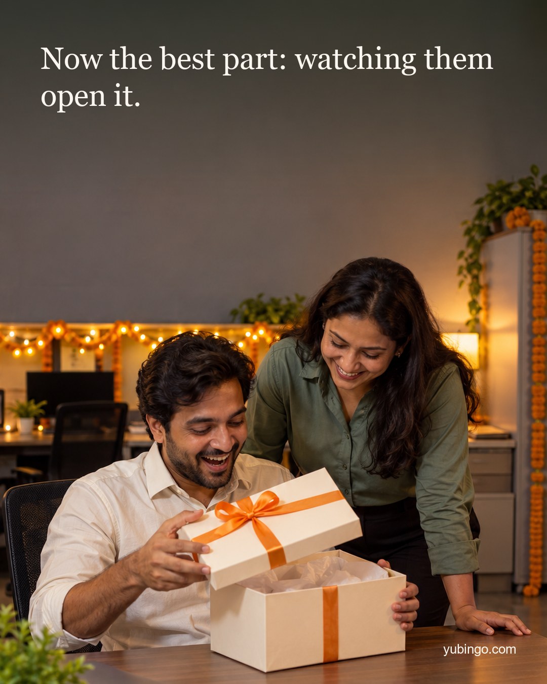 In a festive office with fairy lights, a man opens a gift box at his desk, laughing in surprise, as a colleague leans in. Text: Now the best part: watching them open it.